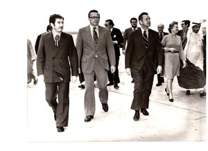 ["The image depicts Dewey Bartlett walking with Fawaz Kudsi, Charge Phillip Griffin, Robert Smith, Nathaniel Howell, Jr., Mrs. Hansen, John Limbert and Rahma Masood across an airstrip"]
