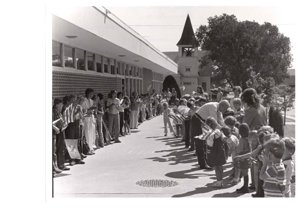 ["The image depicts Dewey Bartlett shaking hands with many school children at the Medford Public School"]